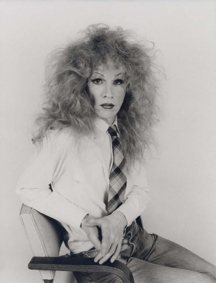 This black and white photograph shows a man wearing a frizzy wig while sitting in a chair.