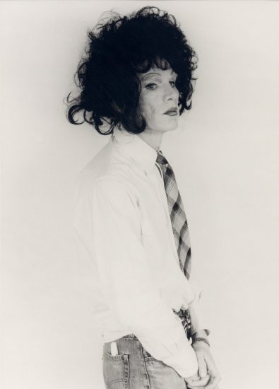 This black and white photograph shows a man standing against a white background wearing a big black wig.