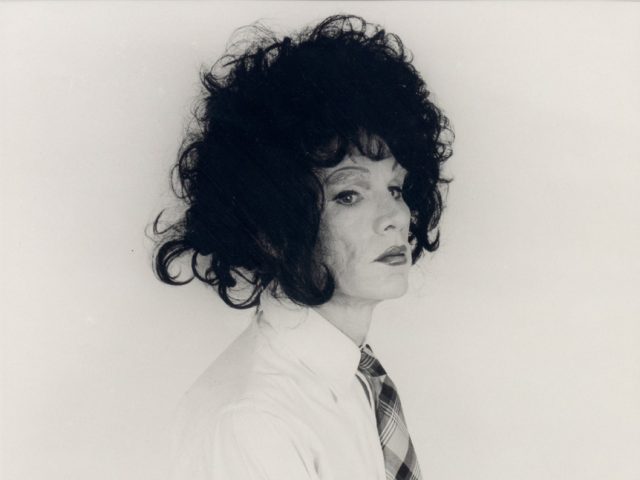 This black and white photograph shows a man standing against a white background wearing a big black wig.