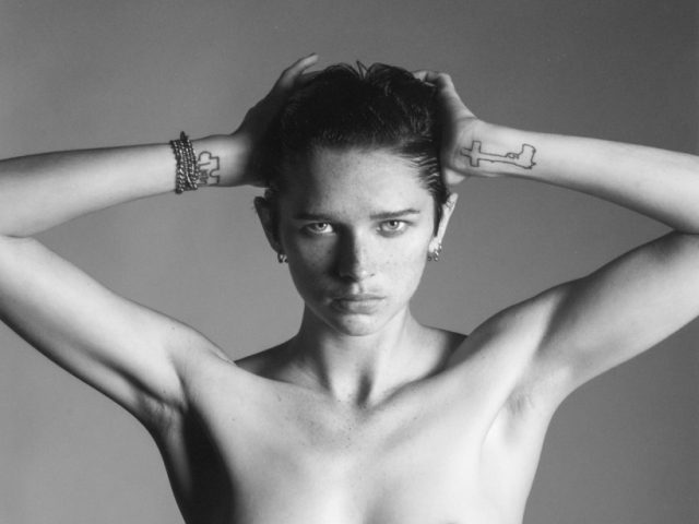 This back and white photograph shows a shirtless woman facing the camera with her hands placed behind her head.