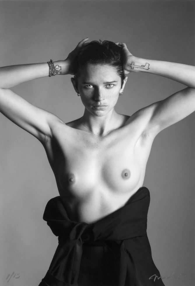 This back and white photograph shows a shirtless woman facing the camera with her hands placed behind her head.