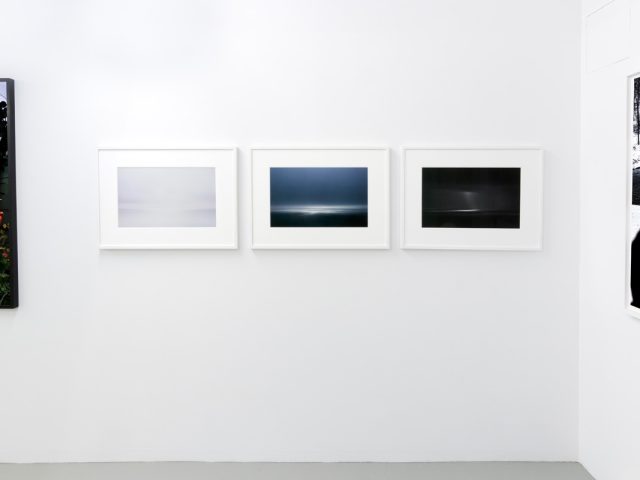 This photograph shows three color photographs of the horizon framed on the walls of a gallery.