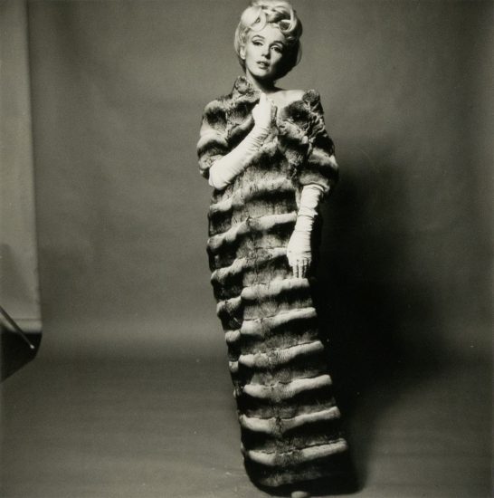 This black and white photograph shows a woman wearing a large, striped fur coat. She stands in front of a grey background and she is wearing a wig.