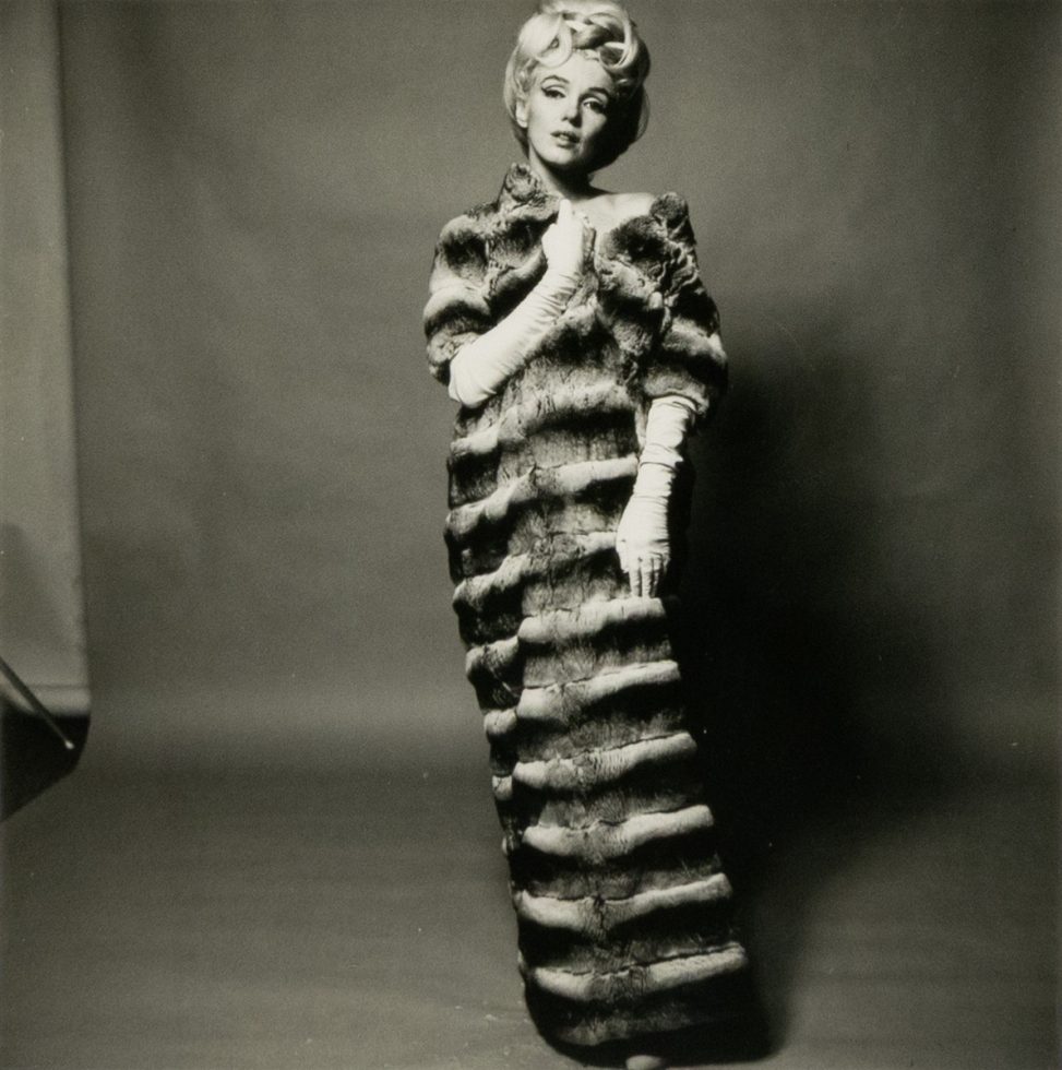 This black and white photograph shows a woman wearing a large, striped fur coat. She stands in front of a grey background and she is wearing a wig.
