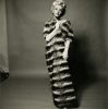 This black and white photograph shows a woman wearing a large, striped fur coat. She stands in front of a grey background and she is wearing a wig.