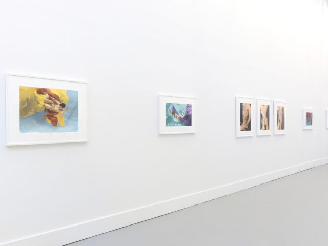 This photograph shows the walls of a gallery with hand painted silver gelatin prints of men floating underwater against floating colorful fabrics.