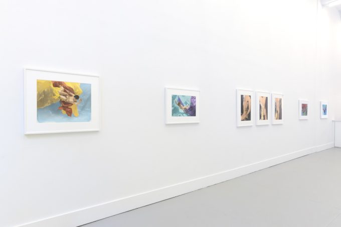 This photograph shows the walls of a gallery with hand painted silver gelatin prints of men floating underwater against floating colorful fabrics.