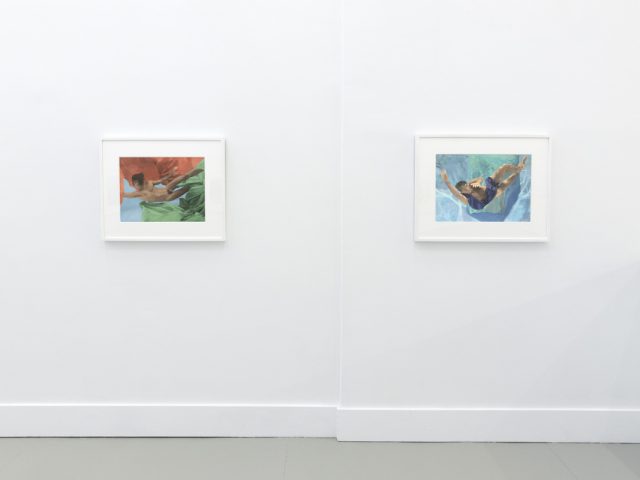 This photograph shows the walls of a gallery with hand painted silver gelatin prints of men floating underwater against floating colorful fabrics.