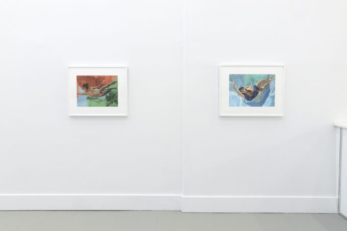 This photograph shows the walls of a gallery with hand painted silver gelatin prints of men floating underwater against floating colorful fabrics.