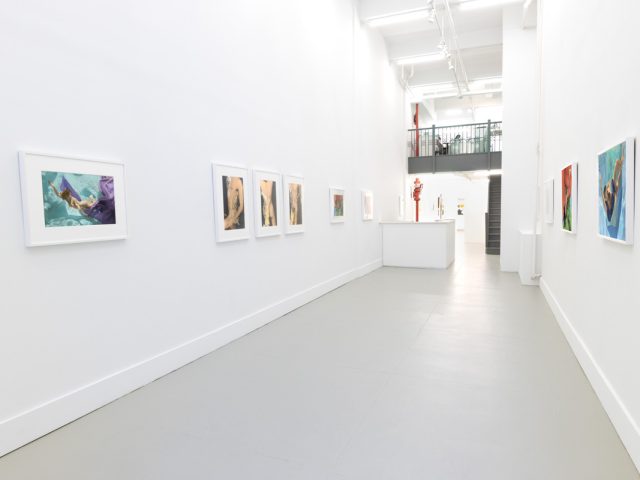 This photograph shows the walls of a gallery with hand painted silver gelatin prints of men floating underwater against floating colorful fabrics.