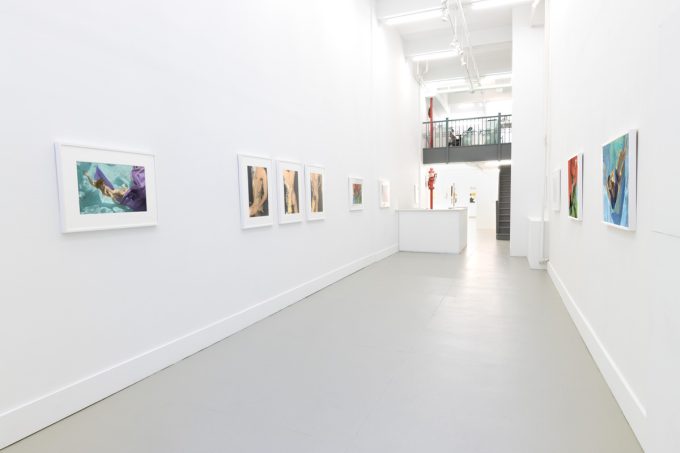 This photograph shows the walls of a gallery with hand painted silver gelatin prints of men floating underwater against floating colorful fabrics.