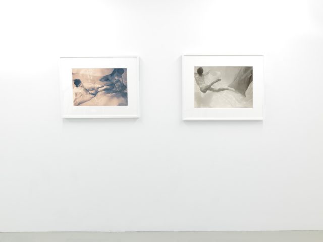 This photograph shows the walls of a gallery with hand painted silver gelatin prints of men floating underwater against floating colorful fabrics.