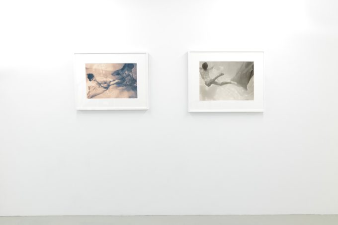 This photograph shows the walls of a gallery with hand painted silver gelatin prints of men floating underwater against floating colorful fabrics.