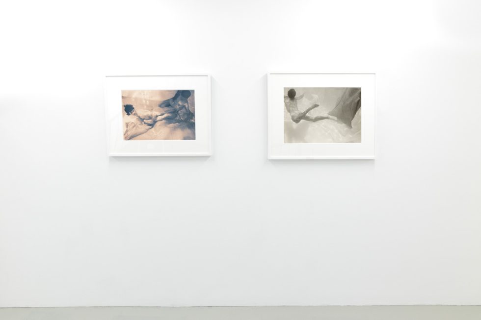 This photograph shows the walls of a gallery with hand painted silver gelatin prints of men floating underwater against floating colorful fabrics.