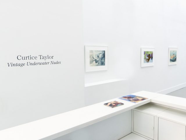 This photograph shows the walls of a gallery with hand painted silver gelatin prints of men floating underwater against floating colorful fabrics.