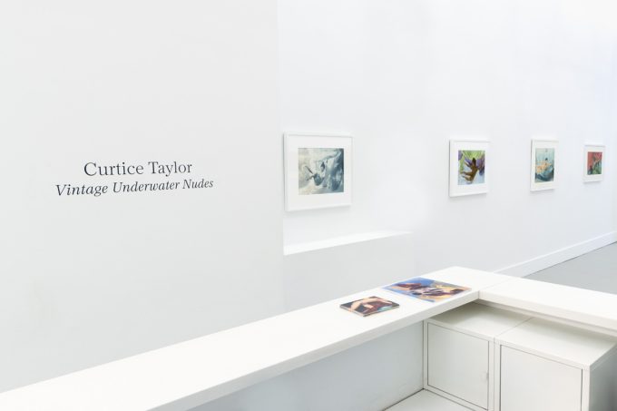 This photograph shows the walls of a gallery with hand painted silver gelatin prints of men floating underwater against floating colorful fabrics.