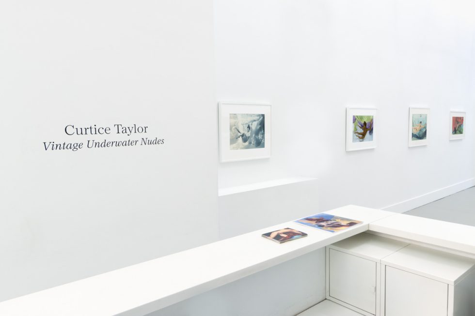 This photograph shows the walls of a gallery with hand painted silver gelatin prints of men floating underwater against floating colorful fabrics.