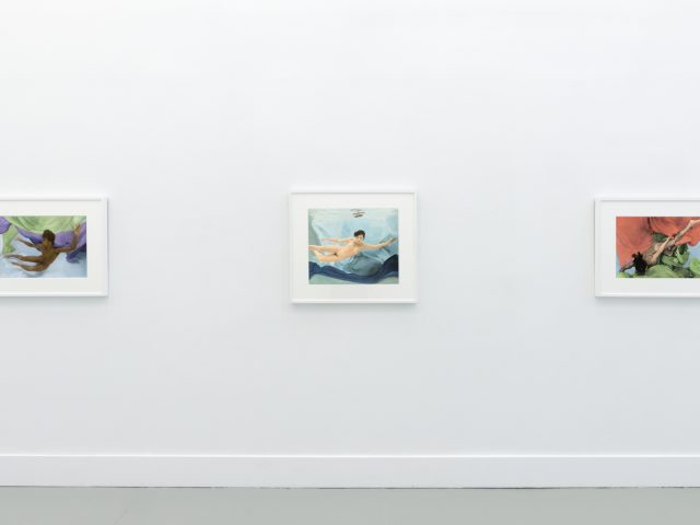 This photograph shows the walls of a gallery with hand painted silver gelatin prints of men floating underwater against floating colorful fabrics.