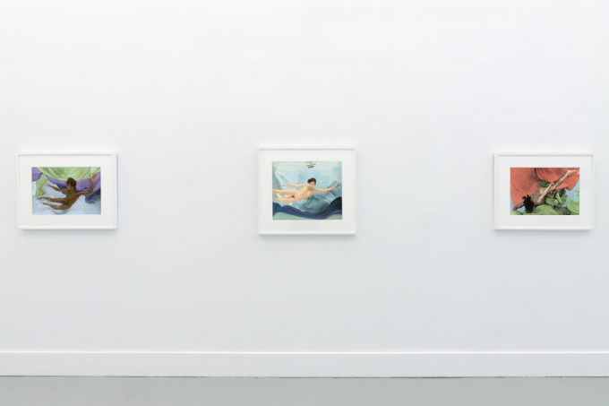 This photograph shows the walls of a gallery with hand painted silver gelatin prints of men floating underwater against floating colorful fabrics.