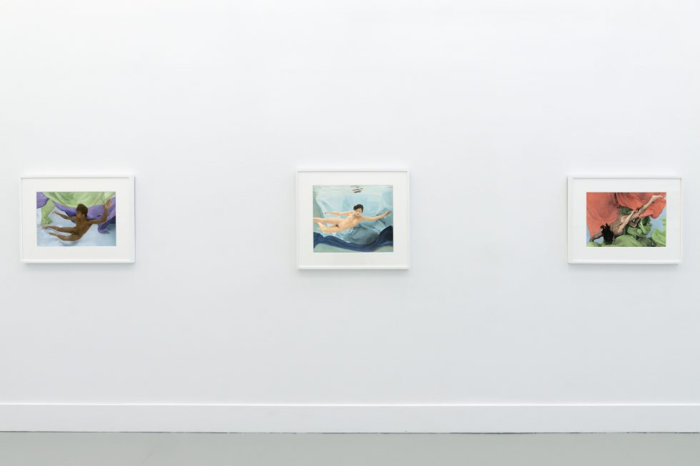 This photograph shows the walls of a gallery with hand painted silver gelatin prints of men floating underwater against floating colorful fabrics.