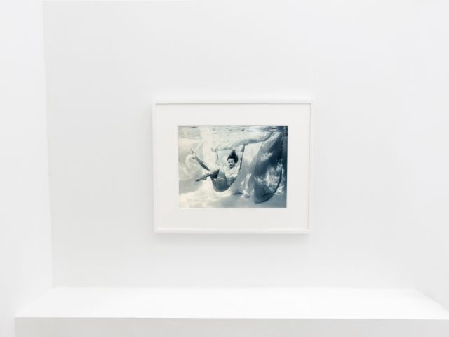 This photograph shows the walls of a gallery with hand painted silver gelatin prints of men floating underwater against floating colorful fabrics.