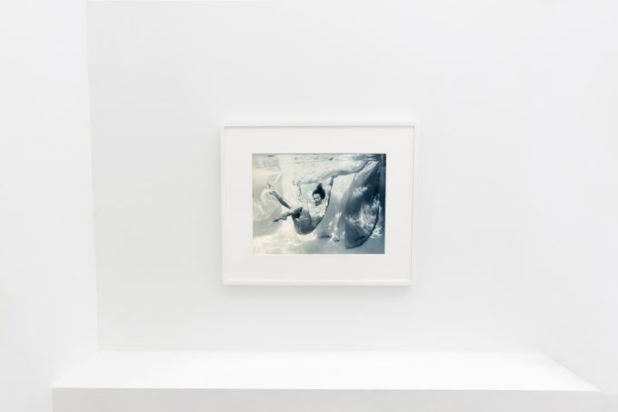 This photograph shows the walls of a gallery with hand painted silver gelatin prints of men floating underwater against floating colorful fabrics.