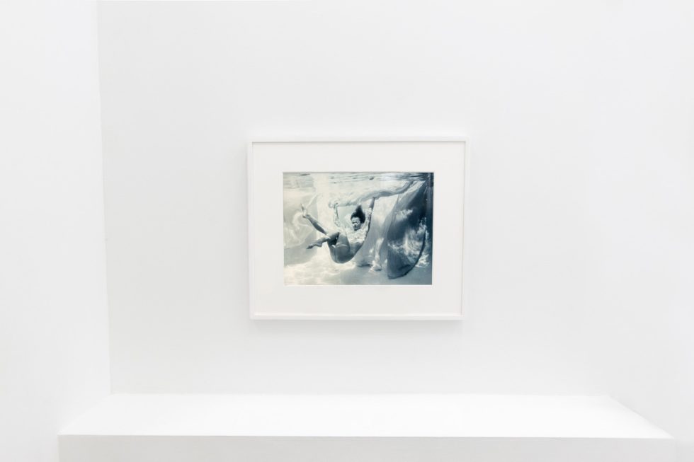 This photograph shows the walls of a gallery with hand painted silver gelatin prints of men floating underwater against floating colorful fabrics.