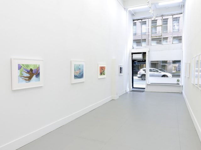 This photograph shows the walls of a gallery with hand painted silver gelatin prints of men floating underwater against floating colorful fabrics.