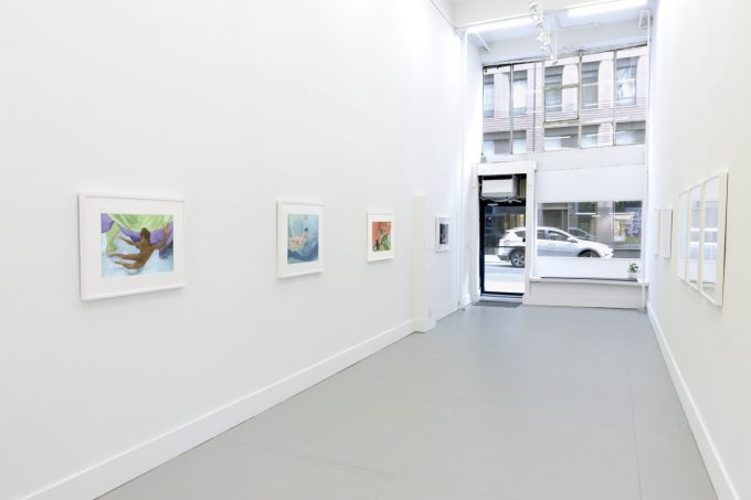 This photograph shows the walls of a gallery with hand painted silver gelatin prints of men floating underwater against floating colorful fabrics.