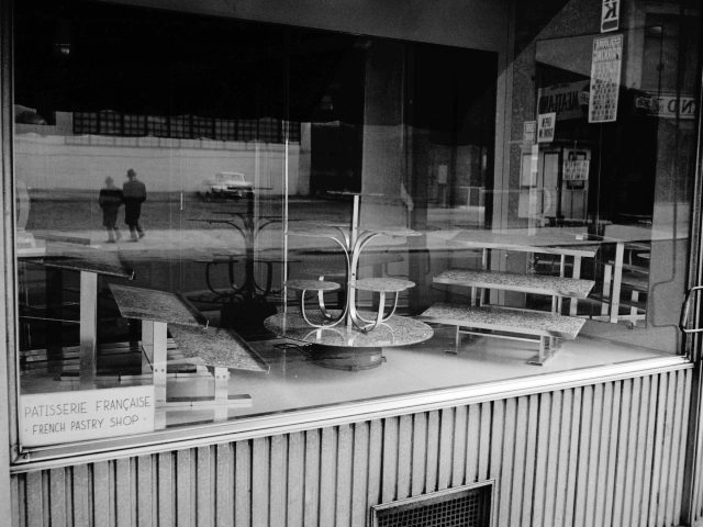 This black and white photograph shows an empty case in a bakery behind a glass window.