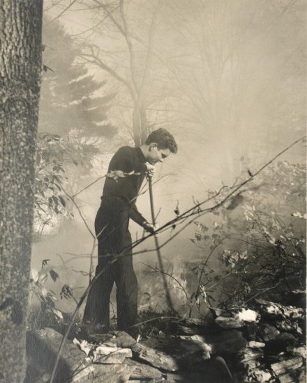 This black and white photograph shows a man standing in a forest while holding a long stick.
