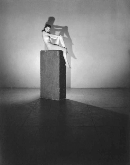 This black and white photograph shows a woman sitting on a column with her hands over her knee. She is naked.