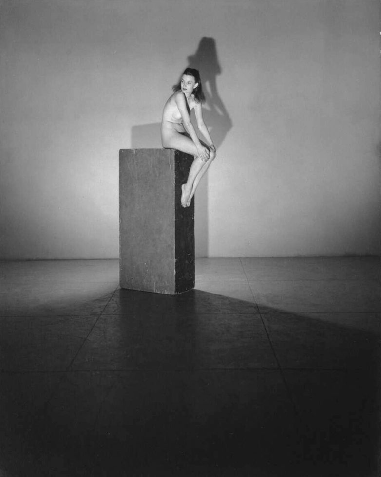 This black and white photograph shows a woman sitting on a column. She is naked.