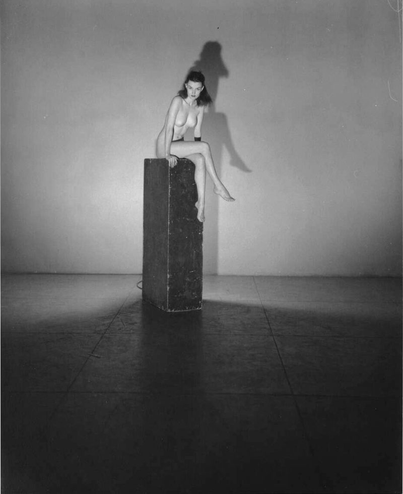 This black and white photograph shows a naked woman sitting on a column with her legs crossed.