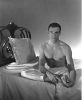 This black and white photograph shows a man sitting on a bed with his foot over his thigh.