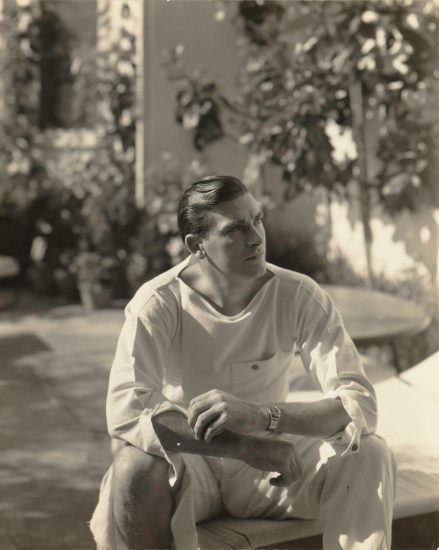 This black and white photograph shows man sitting on a fence and looking off into the distance.