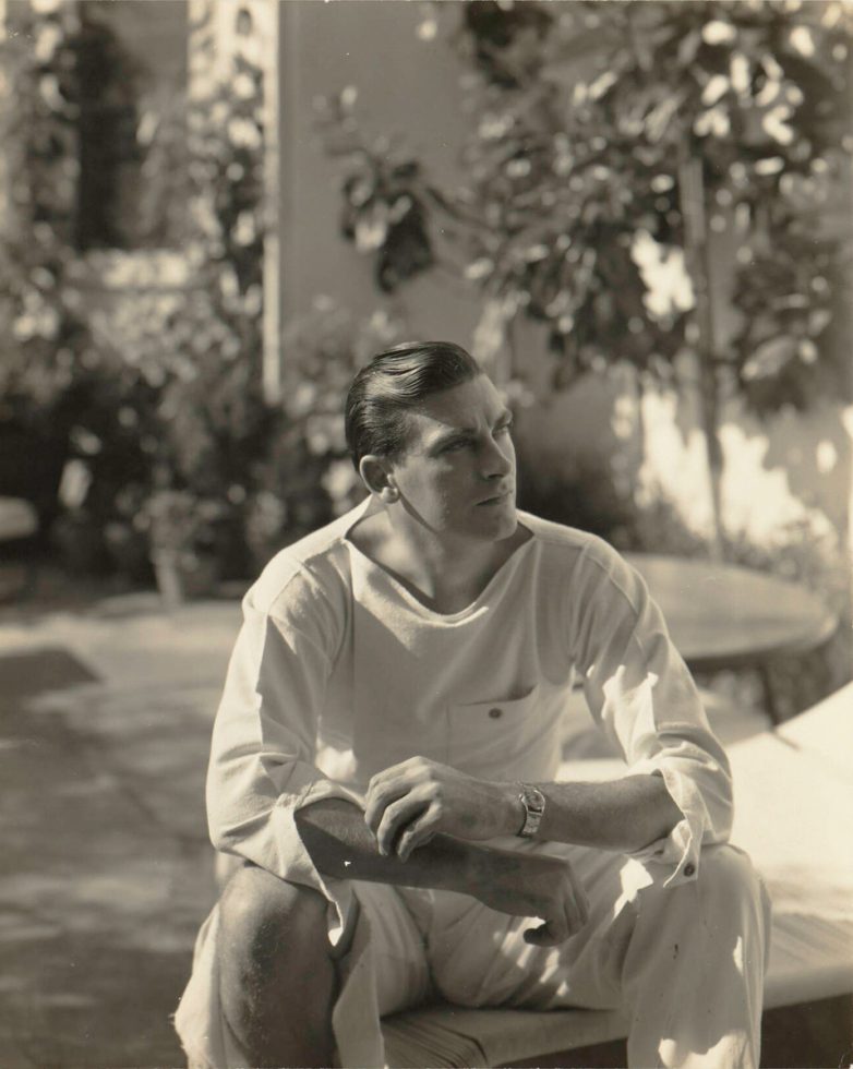This black and white photograph shows man sitting on a fence and looking off into the distance.