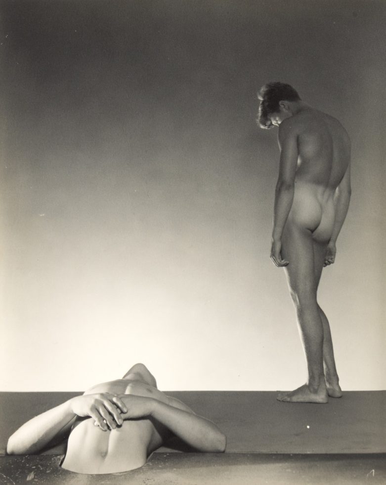 This black and white photograph shows two men against a white background. One is lying down and the other stands over top of him.