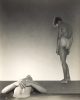 This black and white photograph shows two men against a white background. One is lying down and the other stands over top of him.